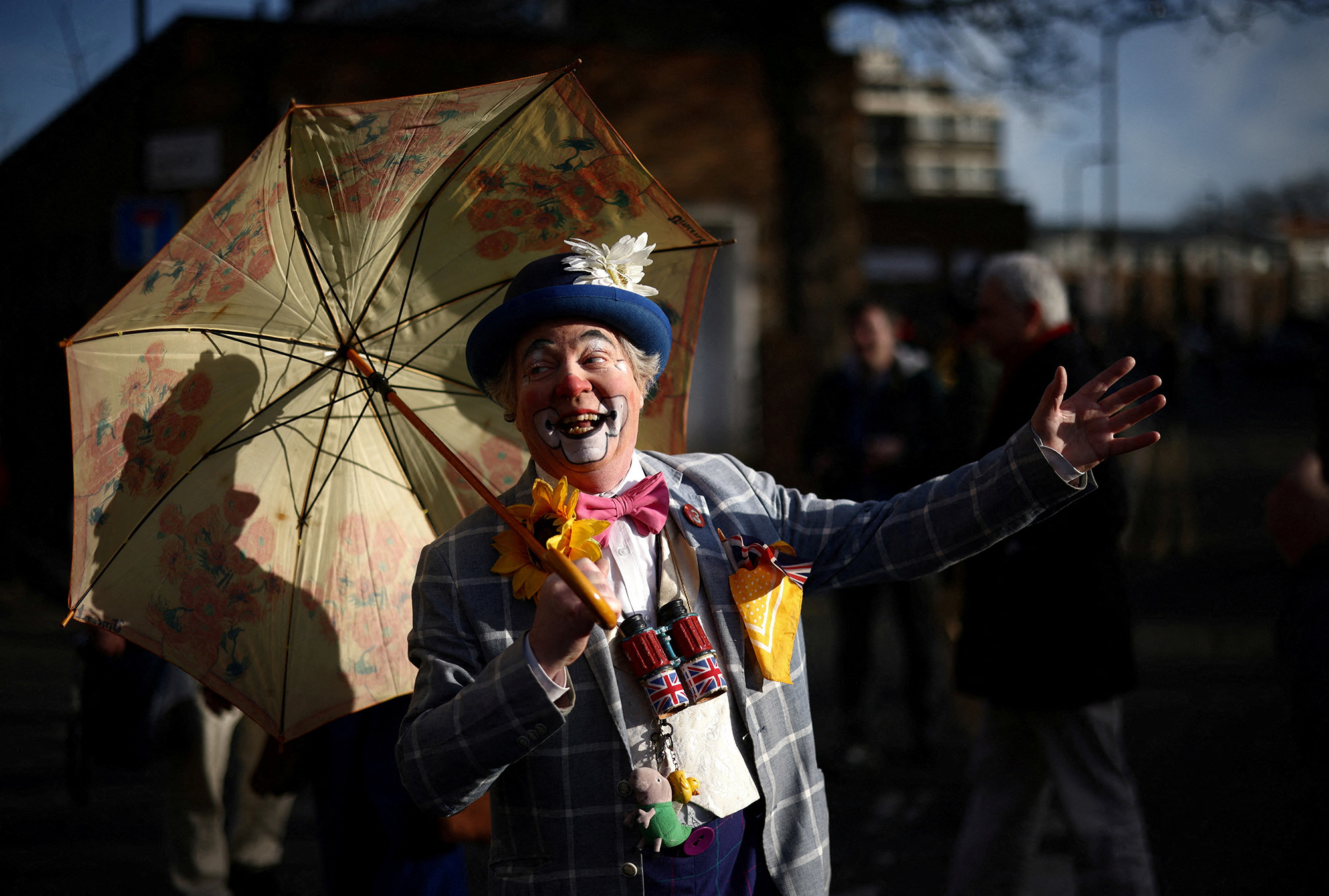 A clown holding a parasol grins and gestures in the shadows