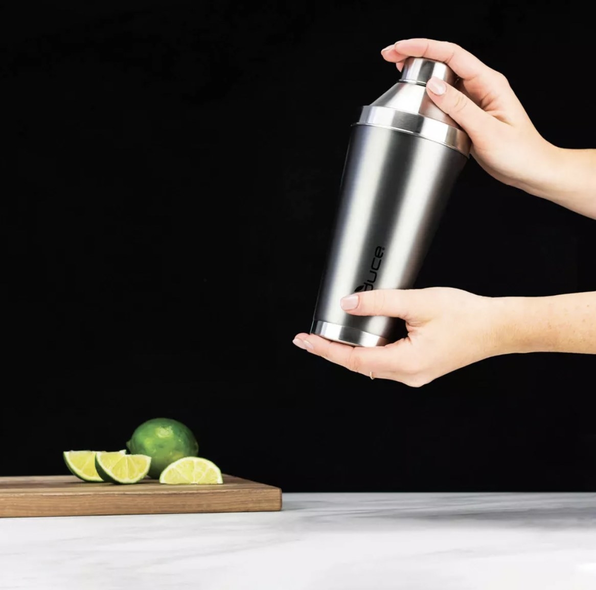 Two hands holding a silver cocktail shaker next to a board with limes on it