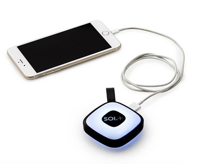 An iPhone plugged into the glowing handbag light via a charging cable. The light is roughly the size of a compact mirror