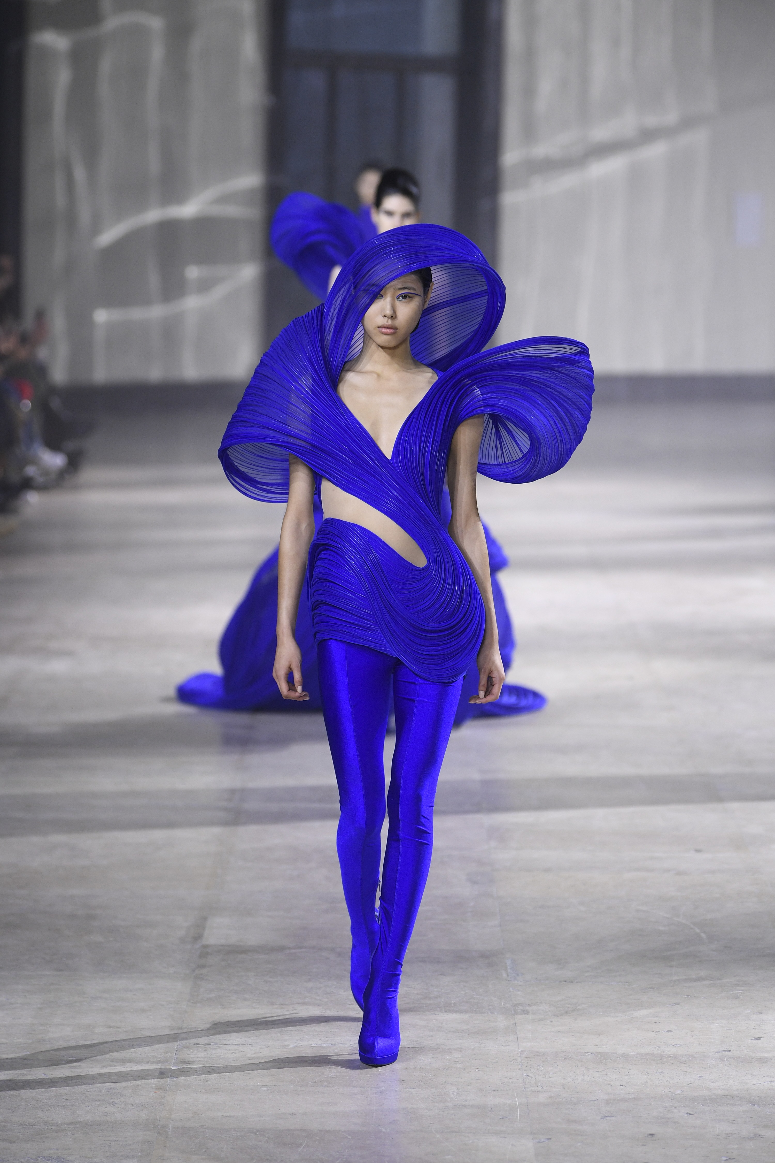 A model walking down the runway in the original piece which features a similar sculpted fabric that sits over the head. This outfit is a jumpsuit rather than a dress