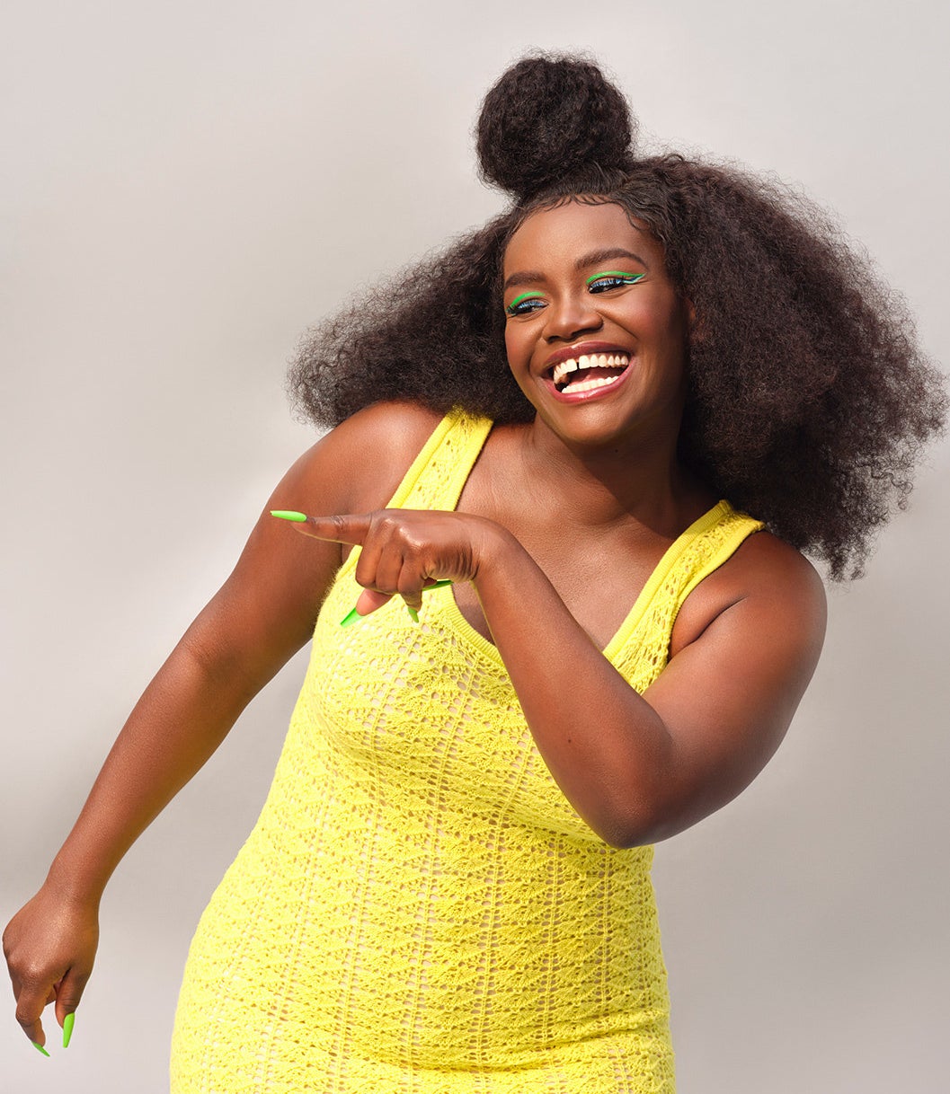 Shoniqua Shandai poses, smiling, in a yellow knit dress