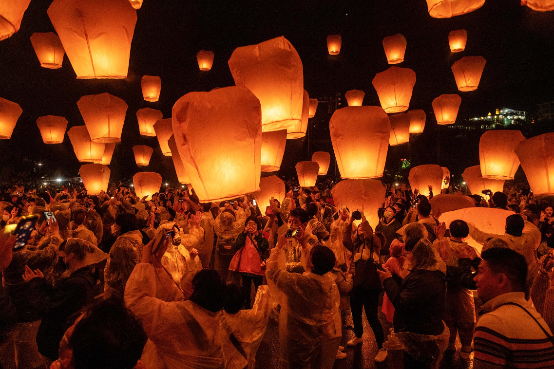 a crowd of people lift glowing orange lanterns up into the night sky