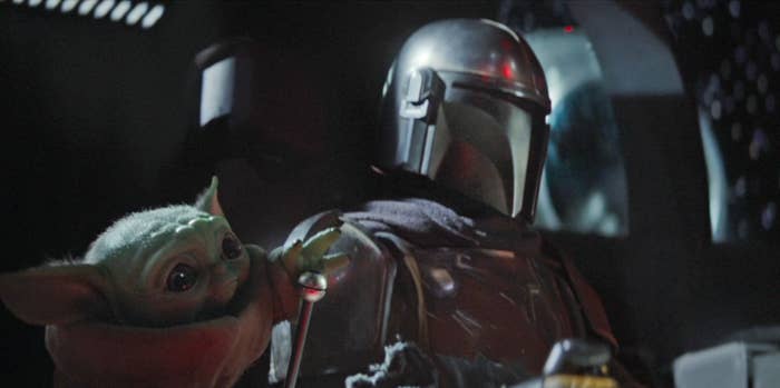 The Child reaches their hand out to pull a lever in Mando's ship while he looks on