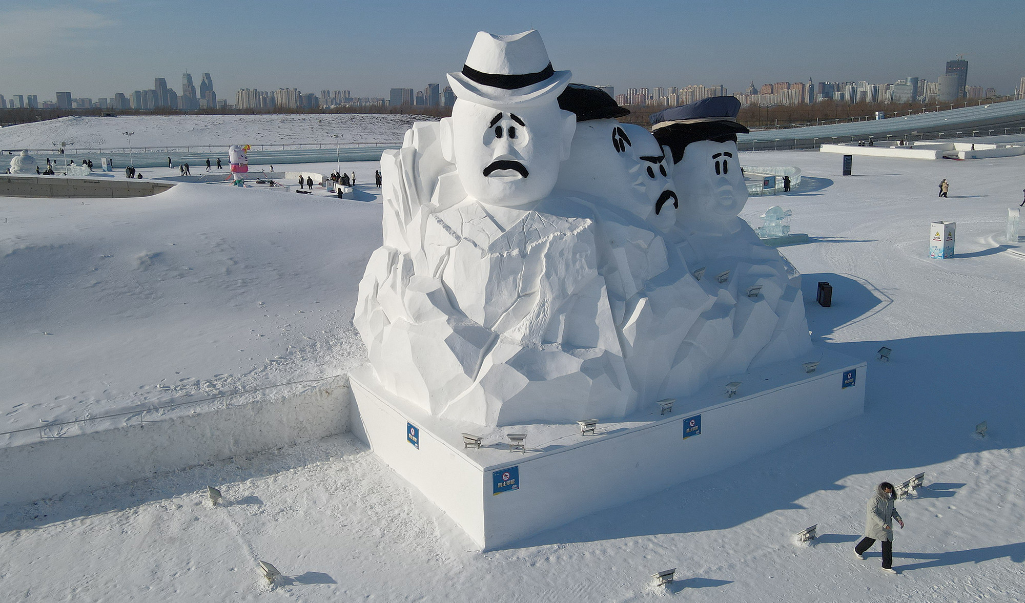 aerial shot of a gigantic snow scultpure with a bunch of cartoon frowny faces; the one directly facing the camera is in a fedora