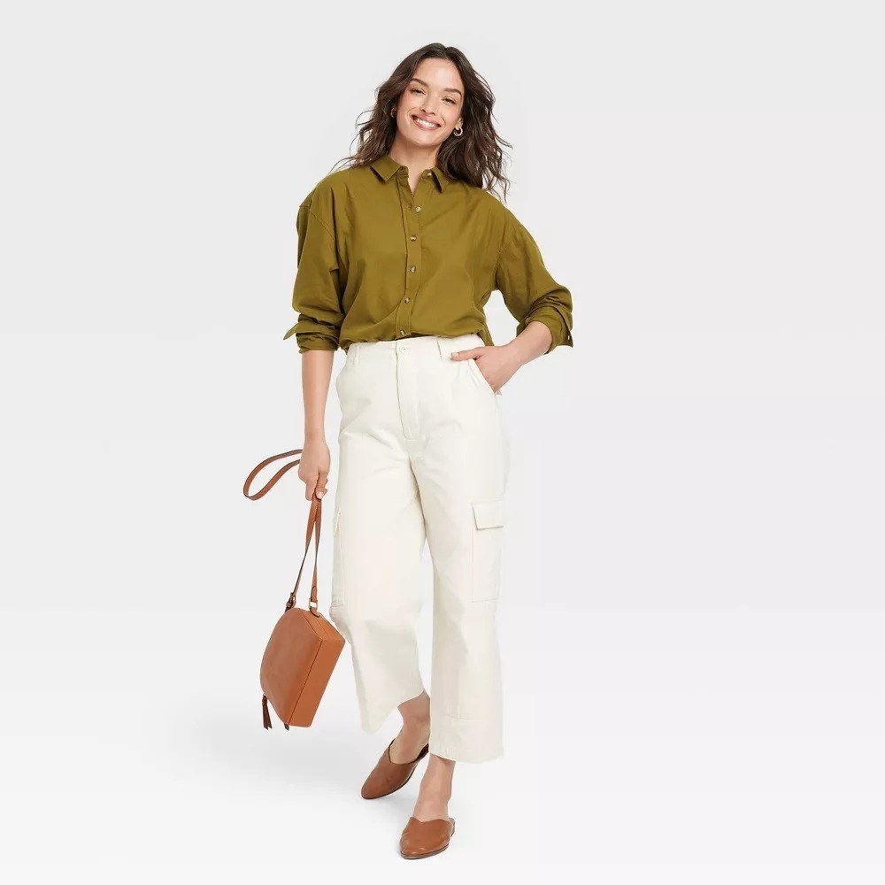 A model wearing the button-down shirt in green while wearing white pants, beige mules and a matching beige purse.