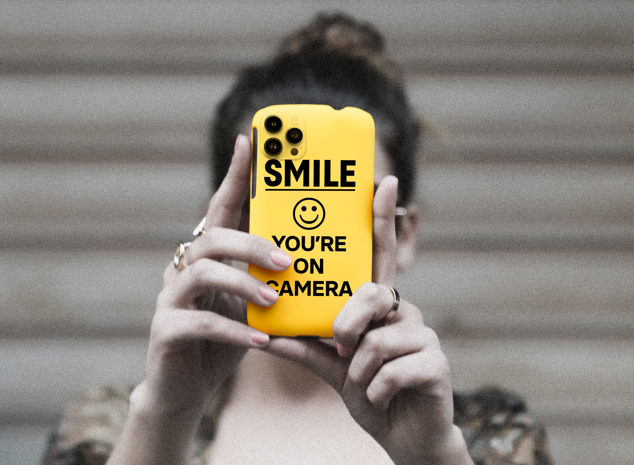A woman holds a phone that resembles a sign that says &quot;Smile, you&#x27;re on camera&quot;