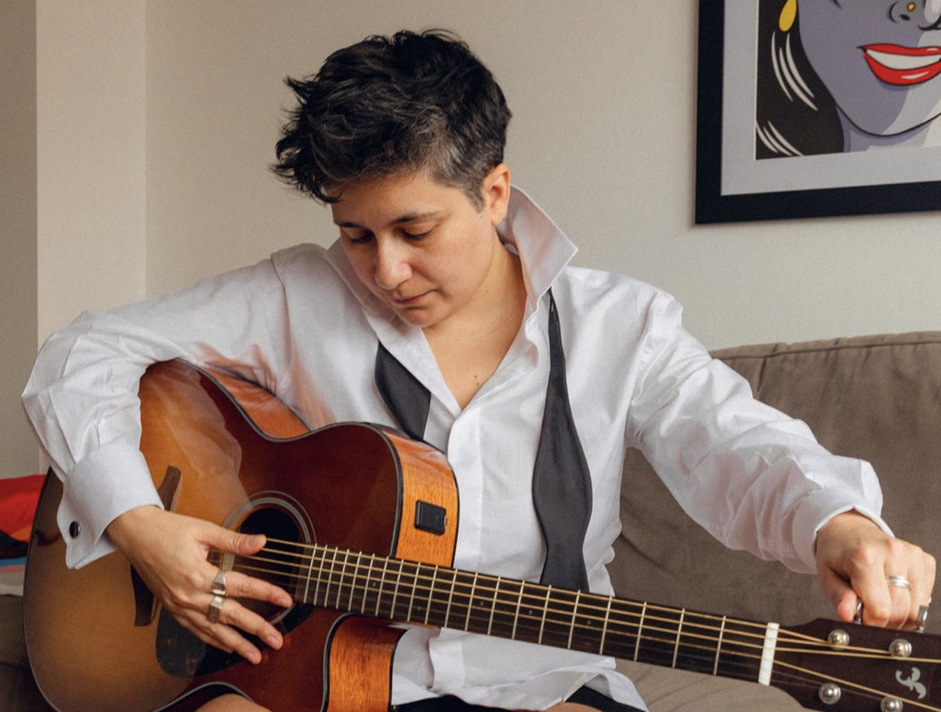 A transmasc person in a button down and undone bow tie playing acoustic guitar