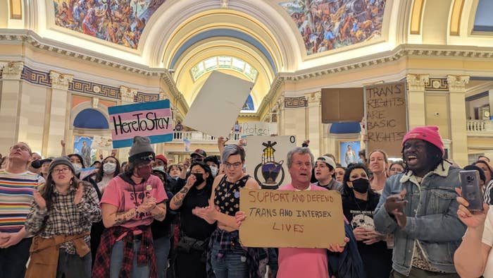 Dozens of trans people hold up signs reading &quot;here to stay&quot; over a handpainted trans flag, and &quot;support and defend trans and intersex lives&quot; and &quot;trans rights are human rights&quot; on pieces of cardboard at a rally at the Oklahoma state capitol