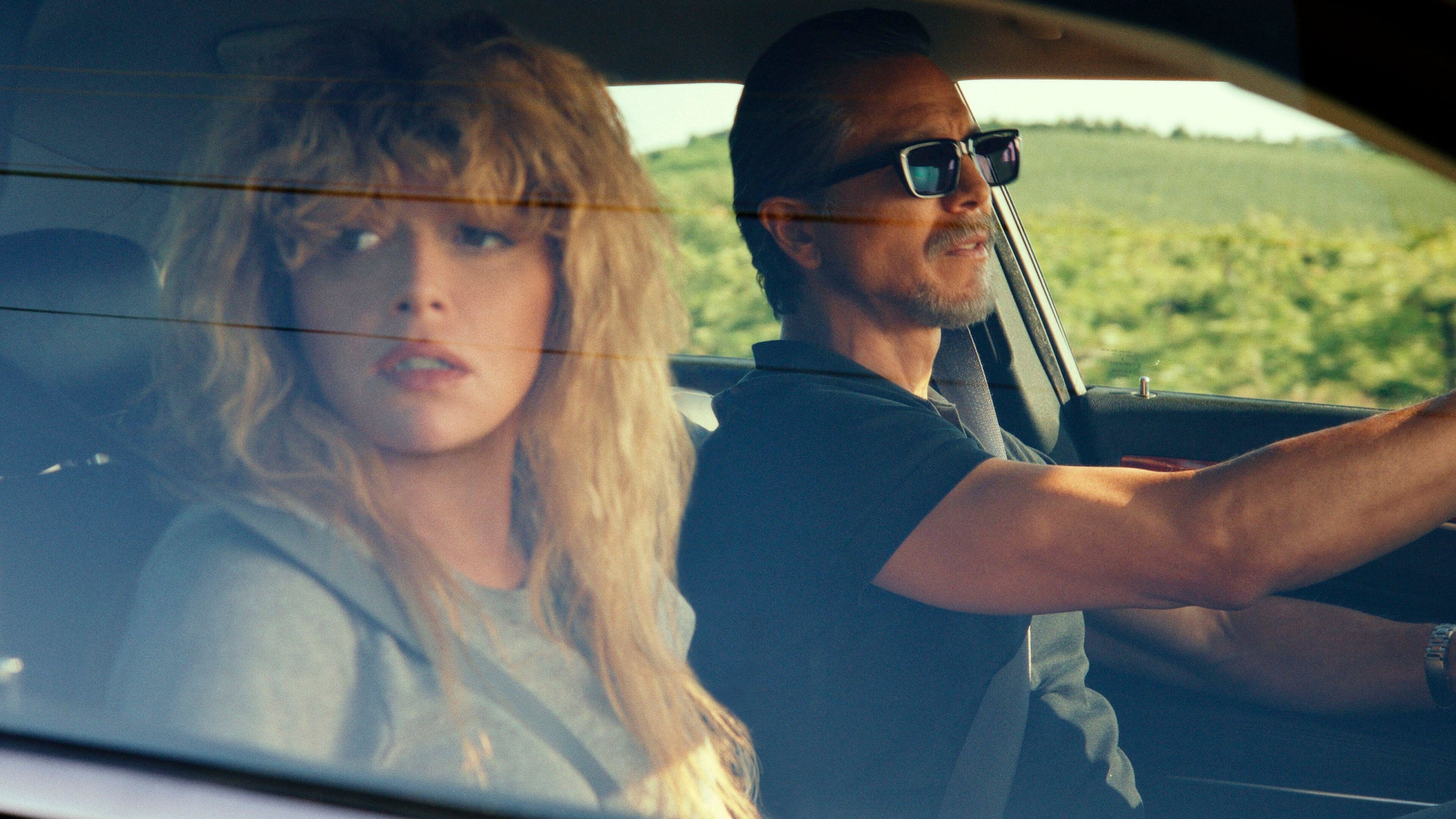 A concerned blonde woman rides next to an intense bearded man driving a car