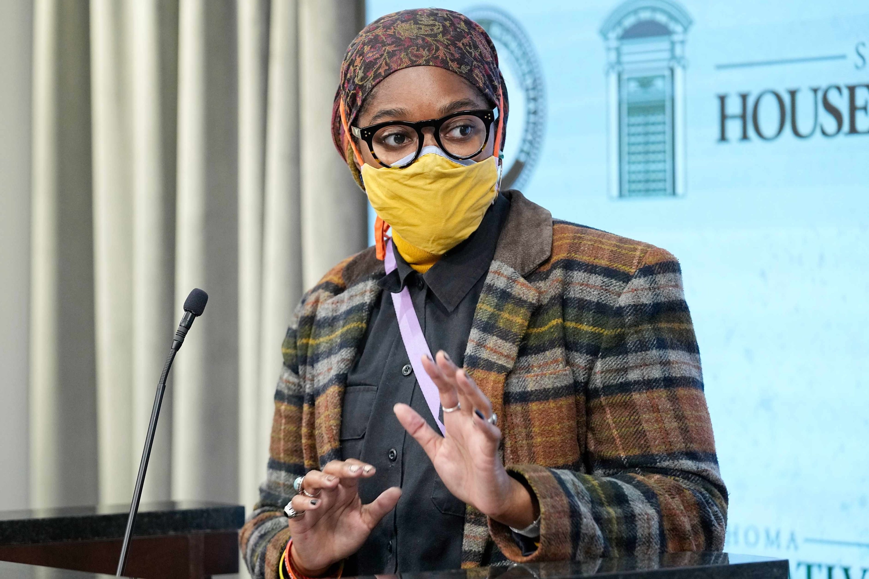 A person stands behind a lectern with a microphone, gesturing with their hands and wearing a yellow face mask