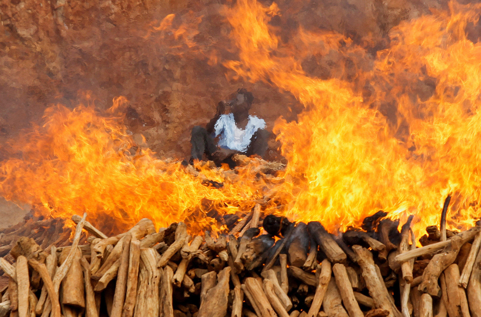 a man sits in the middle of a gigantic wood fire, his image is blurry due to the smoke