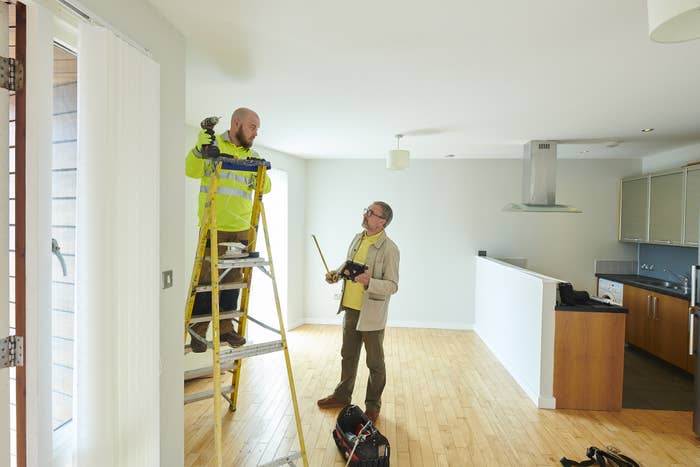 A person stands on a ladder inside of a dining room while another contractor looks on