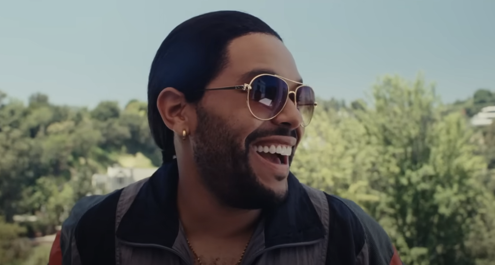 A closeup of The Weeknd wearing sunglasses and smiling widely in a scene from the show