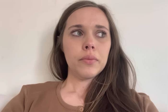 A brunette woman with tears in her eyes sits up against a blank white wall and looks to the left