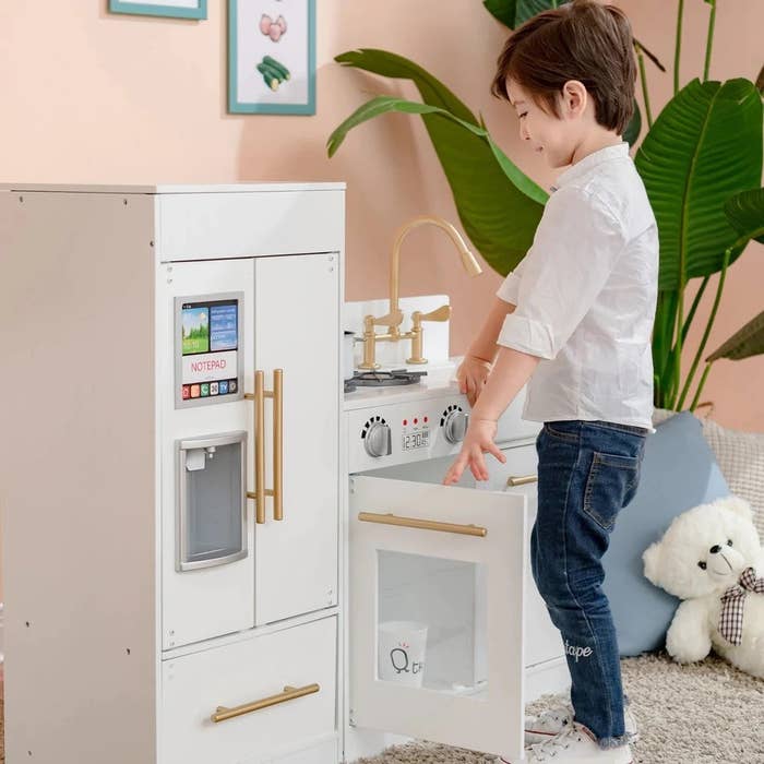 a child model standing in front of the play kitchen set and opening a cabinet