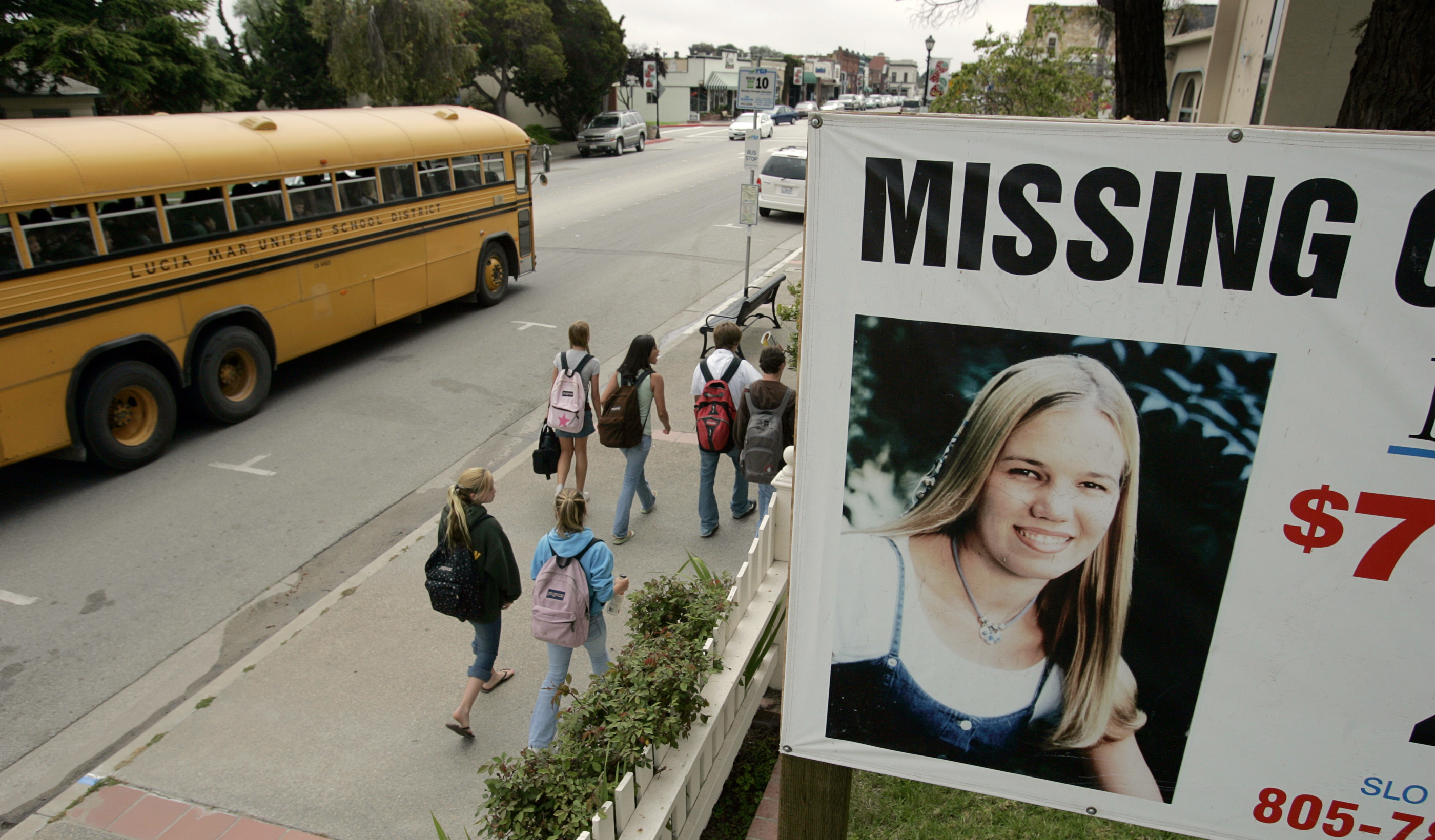 A &quot;Missing&quot; billboard above a sidewalk as young people and a school bus go by