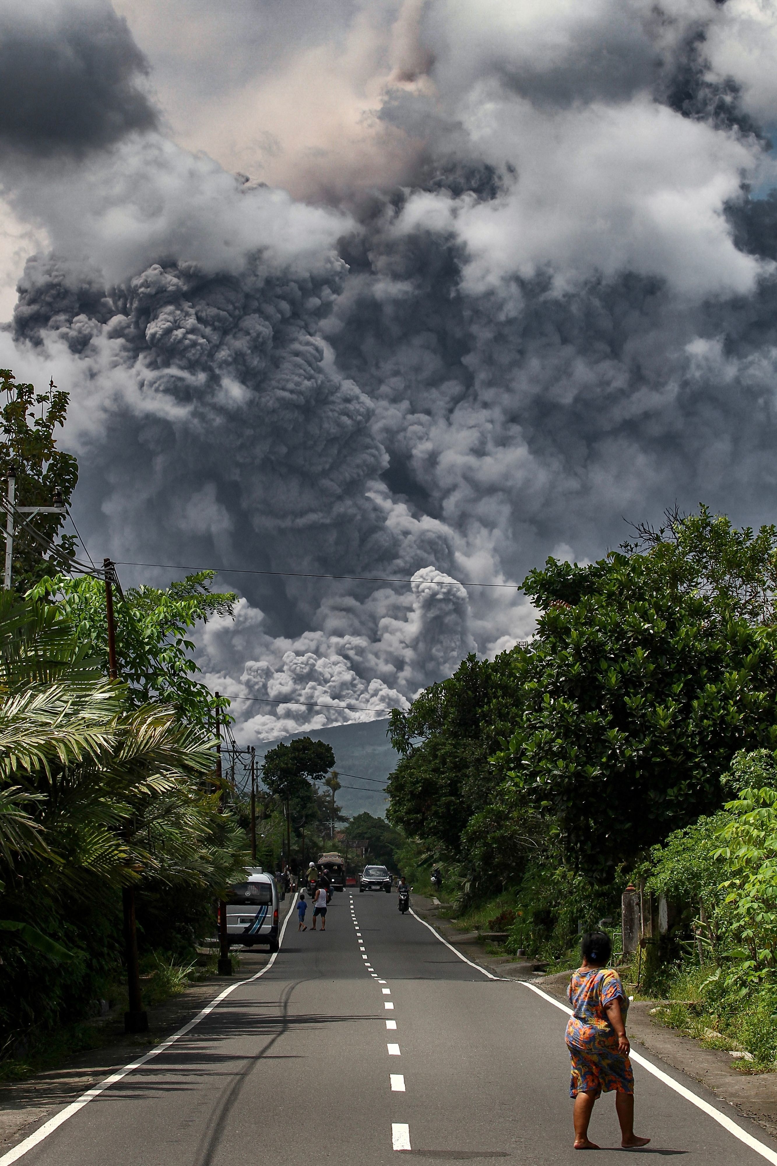 People stand in a road lines with greenery watching pillars of dark smoke obscure the sky