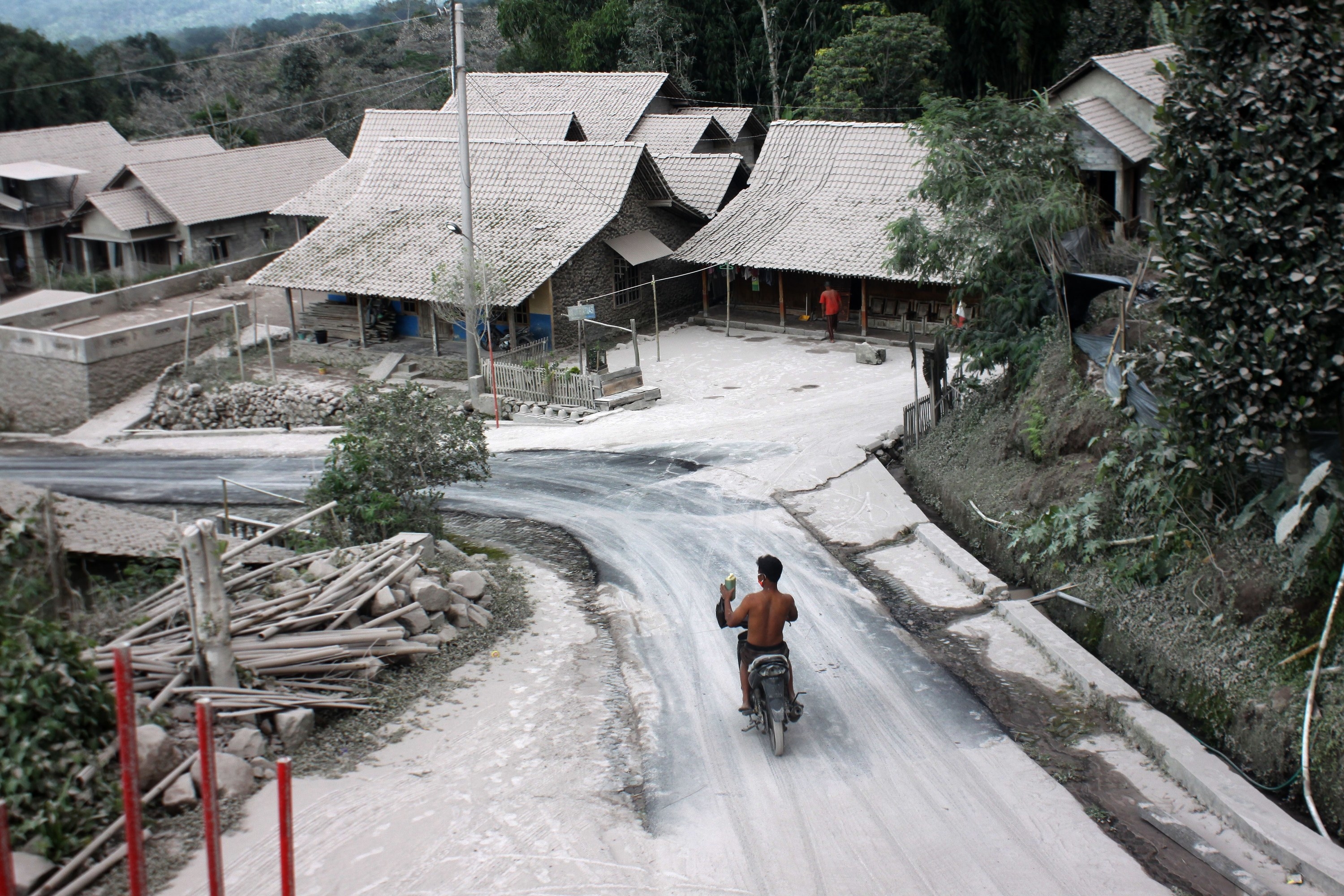 A man on a motorbike rides along a winding road in a residential area with houses covered in a blanket of volcanic ash
