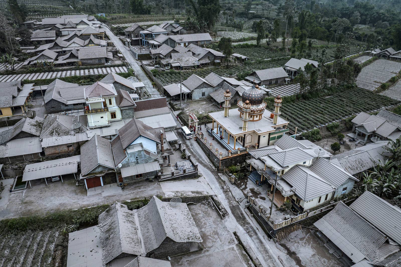 an aerial view of a  village with one to two story houses, a colorful mosque, and fields of crops covered in white ash