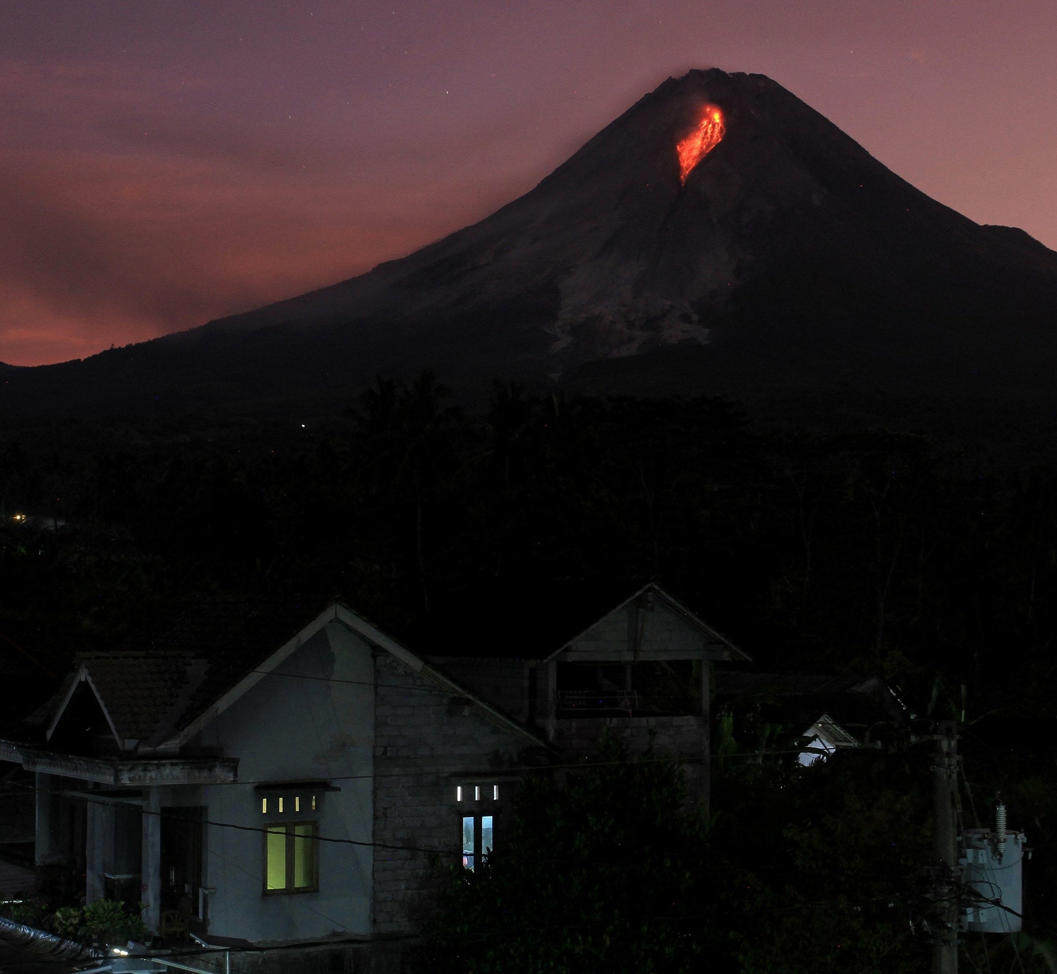 Kaliurang village sitting at the base of Mount Merapi at night as the sun sets, lava pours out of the volcano