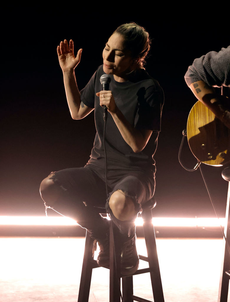 Gaga sitting on a stool onstage holding a microphone with torn pants, T-shirt, and no makeup