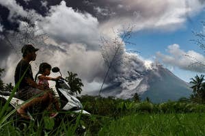 A man and a child sit on a motorcycle in a green field with Mount Merapi in the background spewing thick ash and smoke that darkens the sky