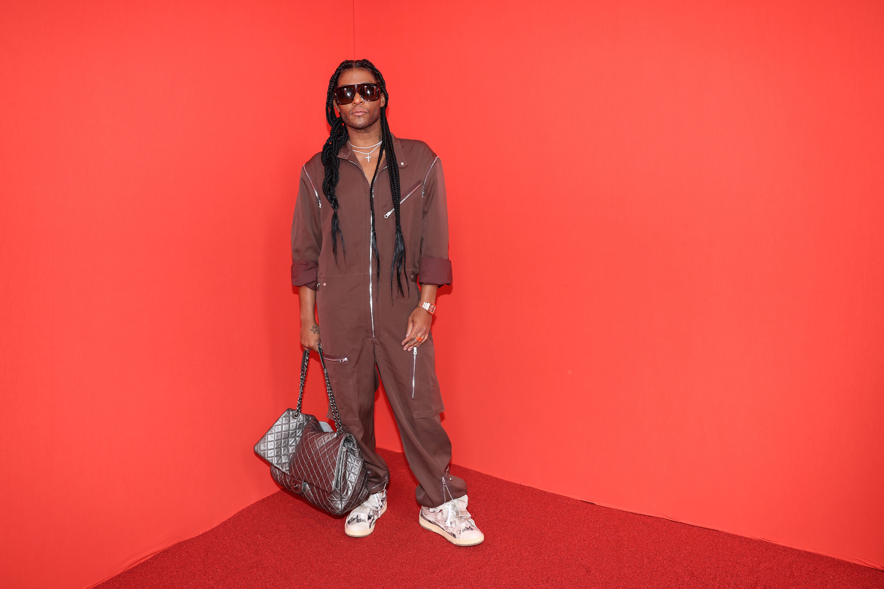 Law Roach wears a burgundy one-piece outfit and carries a high-fashion duffel bag, standing in the corner of a bright red-walled space