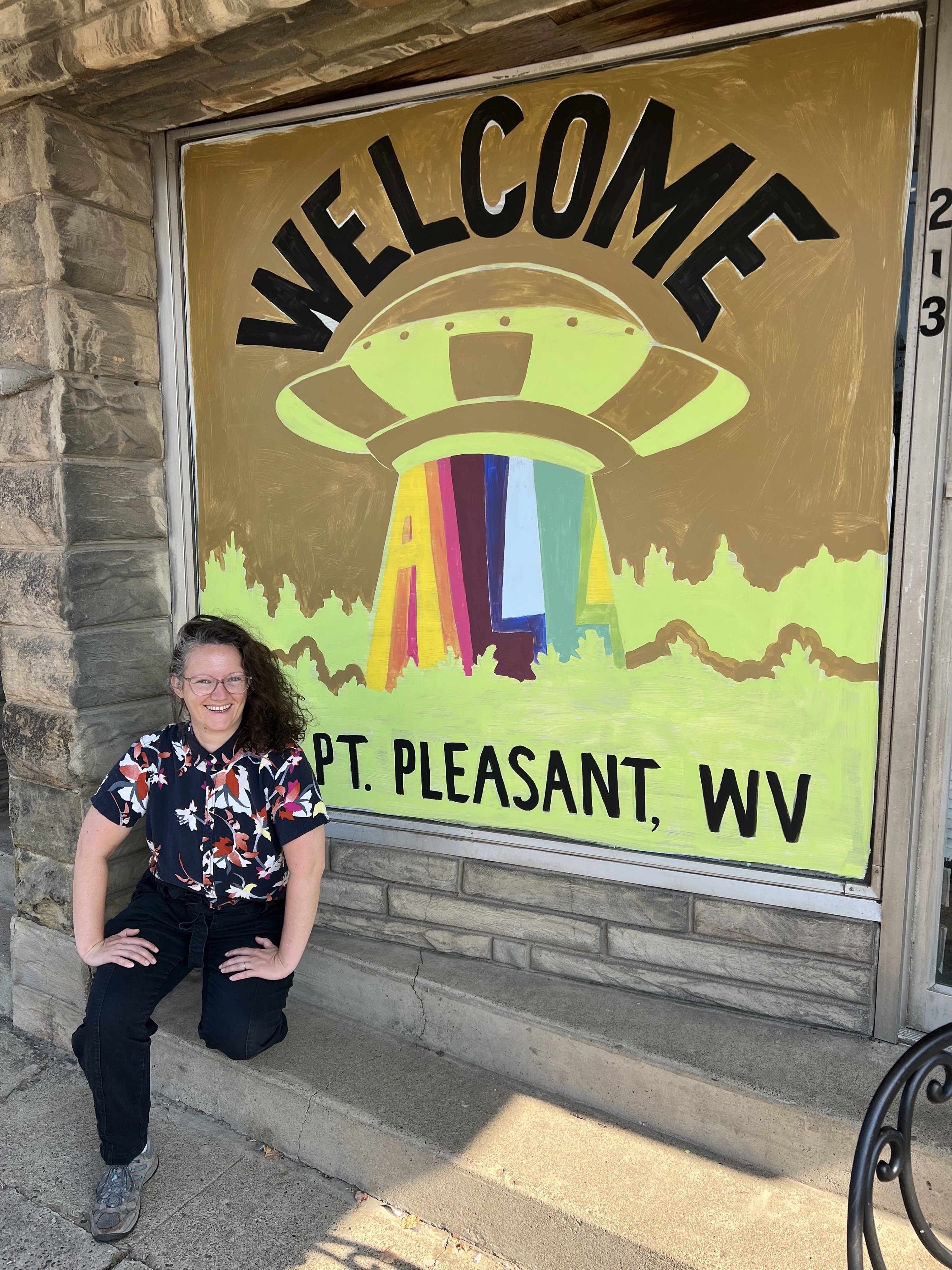 Woman in front of a sign that has a UFO and says "Welcome to Point Pleasant West Virginia"