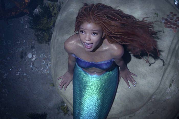 halle bailey as the live-action little mermaid, in a seashell bra and fins, sitting on a rock, singing toward the sky