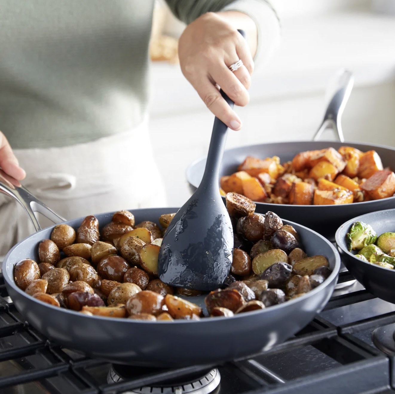 Someone sauteing pan roasted potatoes on stove, with fry pans of sweet potatoes and brussel sprouts on other burners