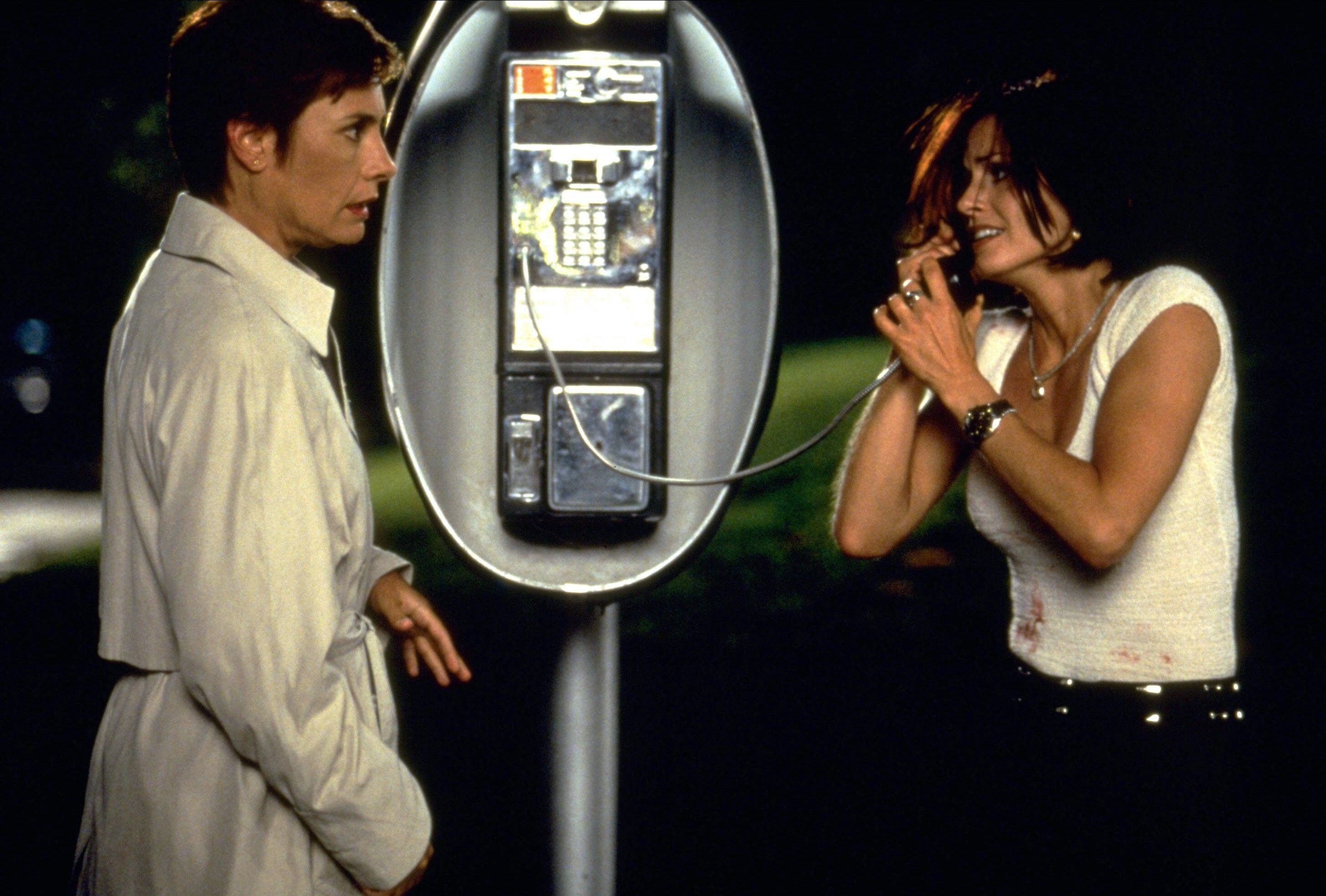 A woman in a trench coat surprises a another woman in a white shirt at a pay phone