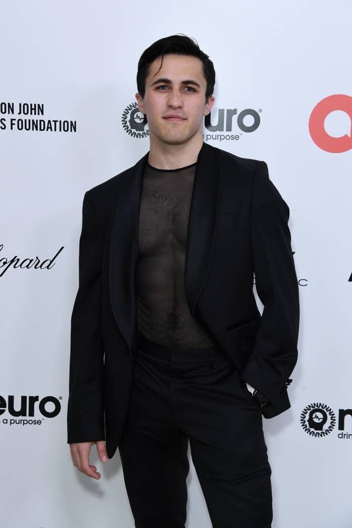 Chris Olsen stands with his hand in his pocket as he poses for photographers at a red carpet event. He&#x27;s wearing a suit and a sheer shirt