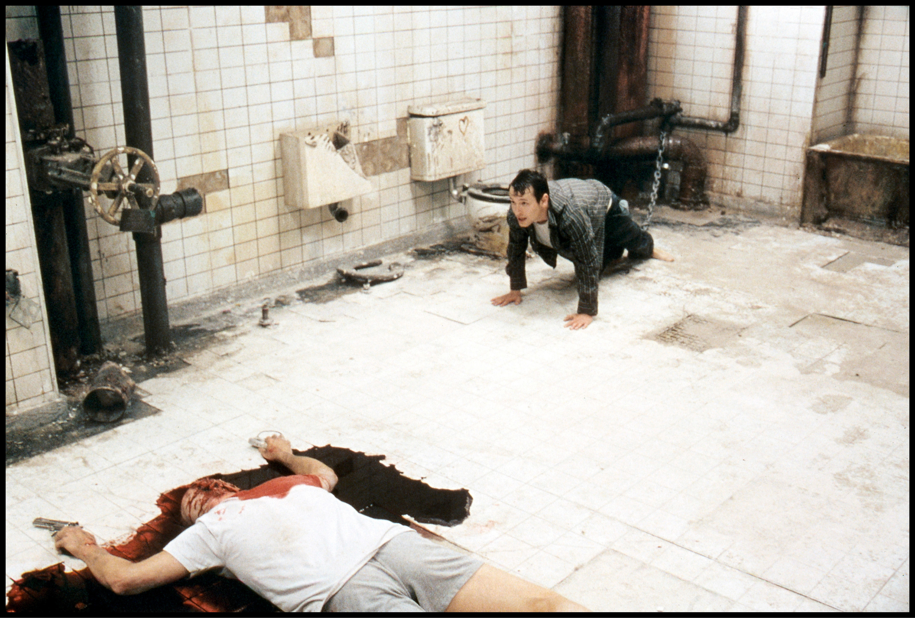A desperate man crawls on a dirty bathroom floor near a victim of an apparent suicide