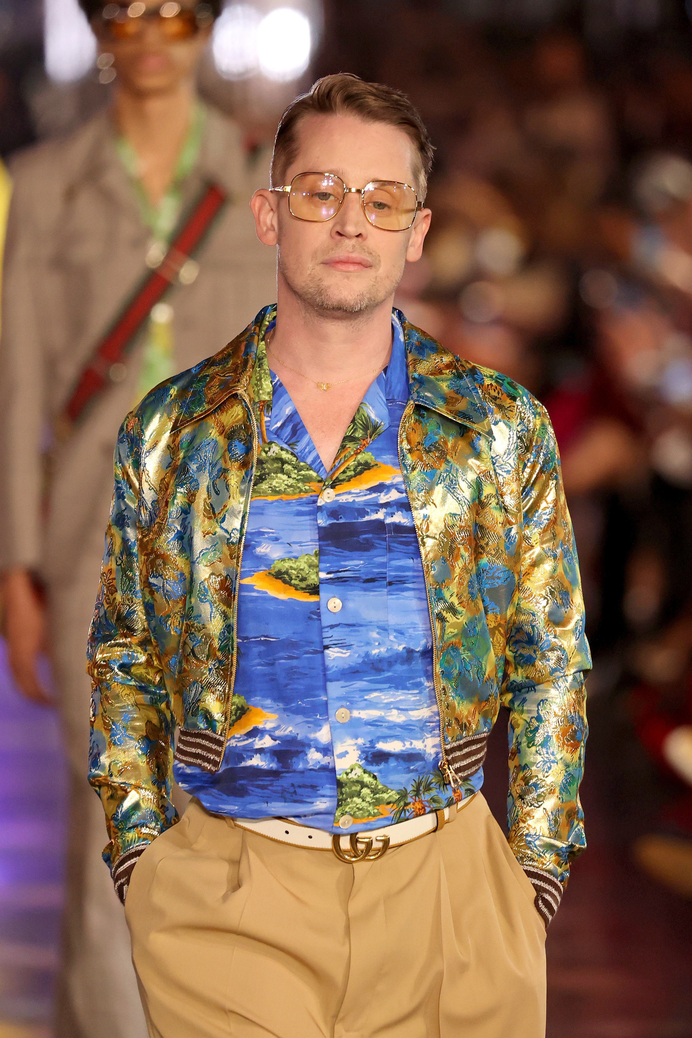 Macaulay on Prada runway, wearing a bomber jacket in the colours of a peacock&#x27;s tail over a blue shirt with the sea and several small islands on it, and camel colored trousers.