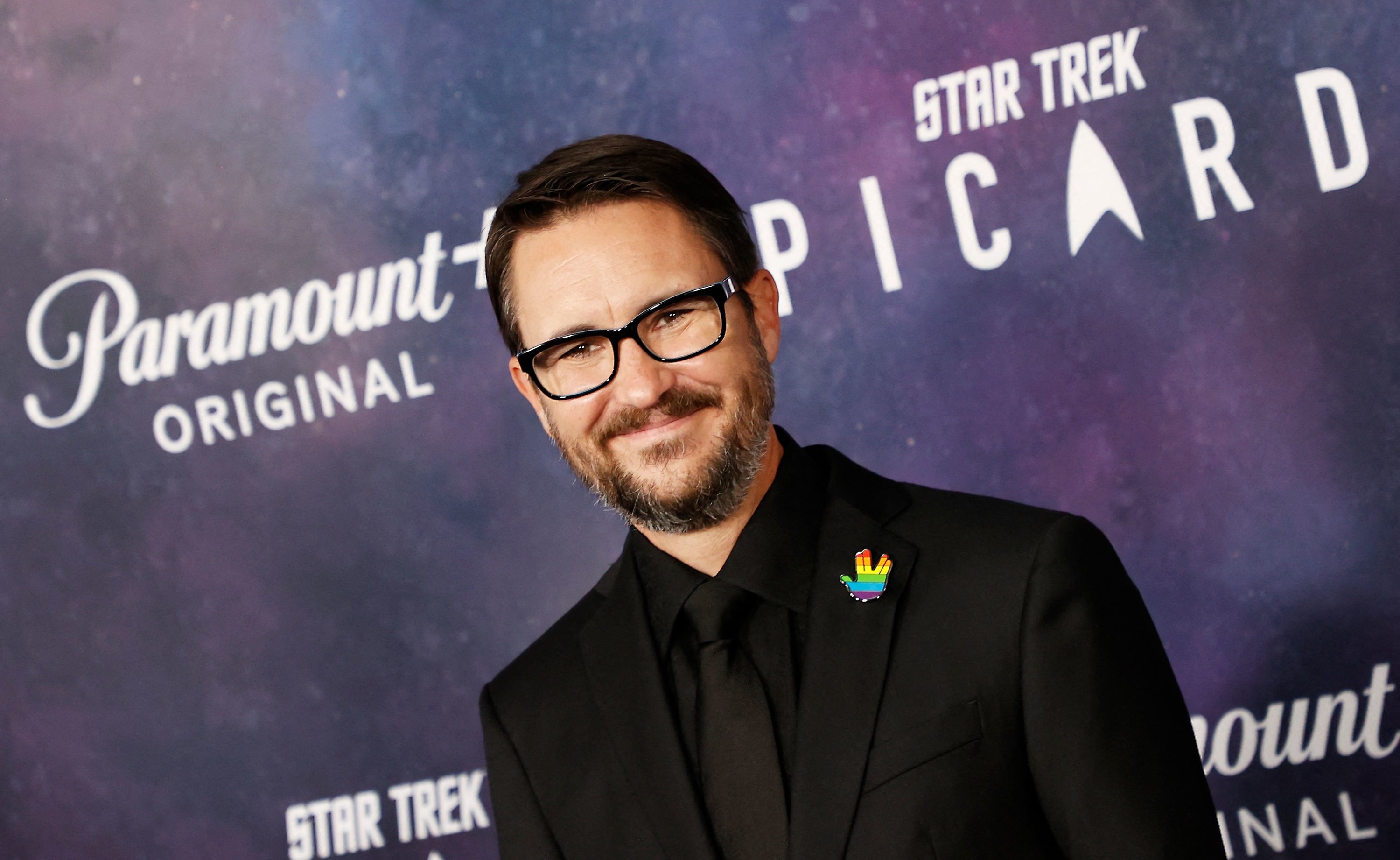 Head and shoulders shot of Wil Wheaton wearing a black suit with an LGBTQ+ Star Trek badge on its lapel