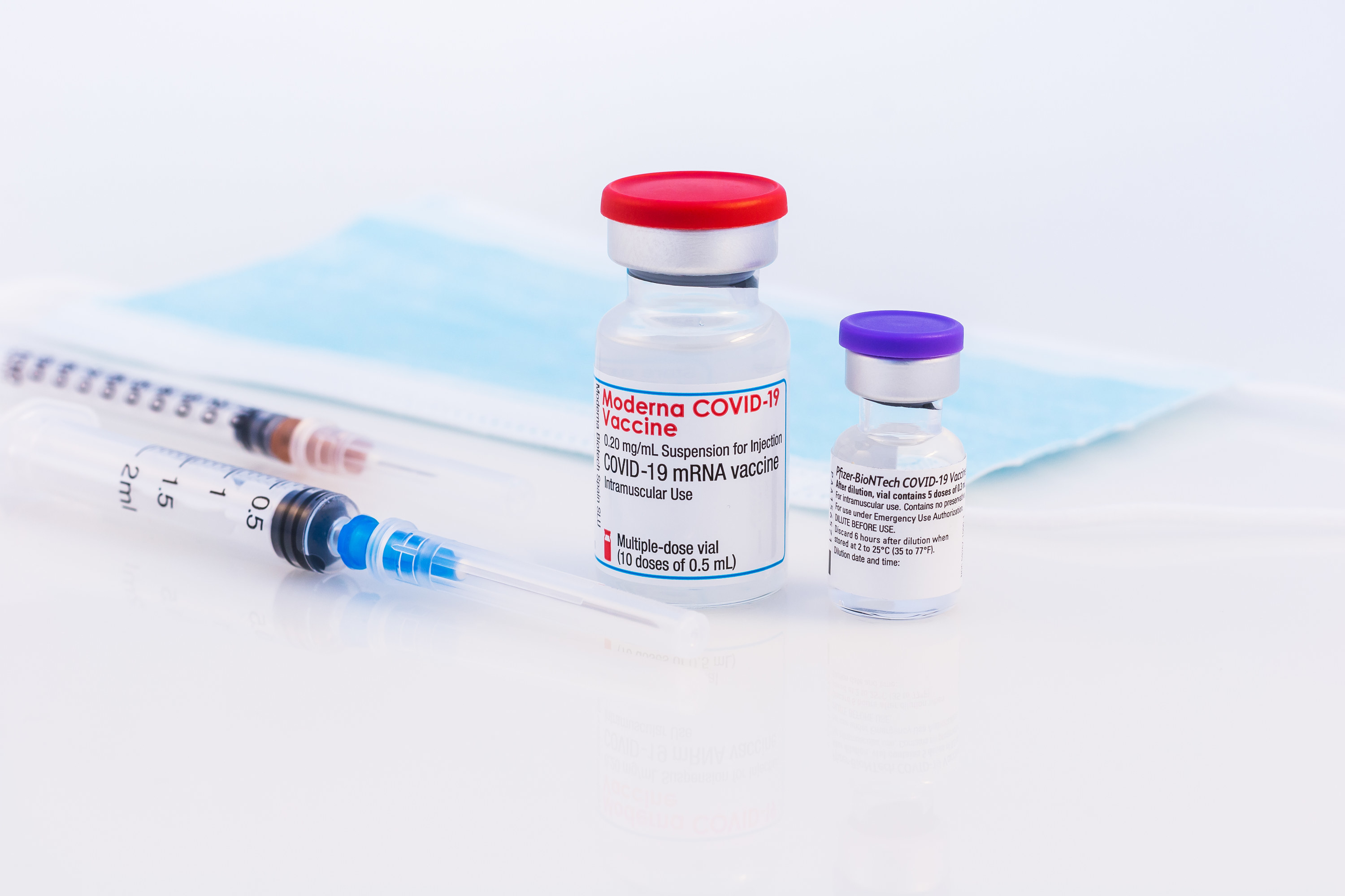 A bottle of the Moderna COVID-19 Vaccine next to two syringes and a surgical face mask