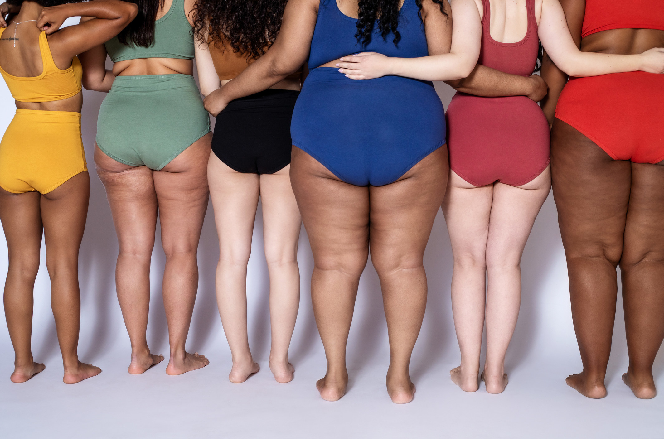 Women of all different shapes and sizes with their backs to the camera in a stock photo. They&#x27;re all wearing sports bras and underwear in various colors