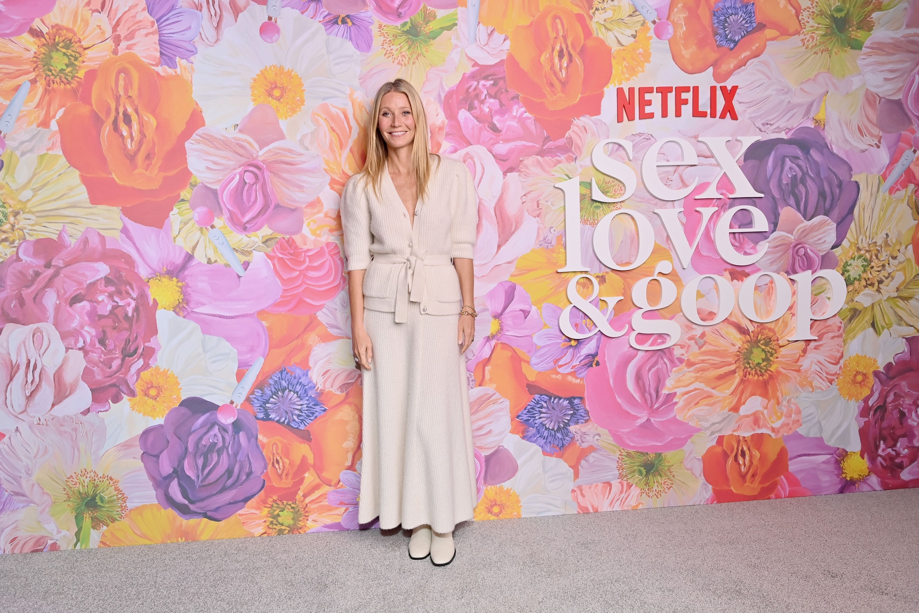 Gwyneth smiles as she stands against a floral backdrop for an event for Sex, Love &amp; Goop
