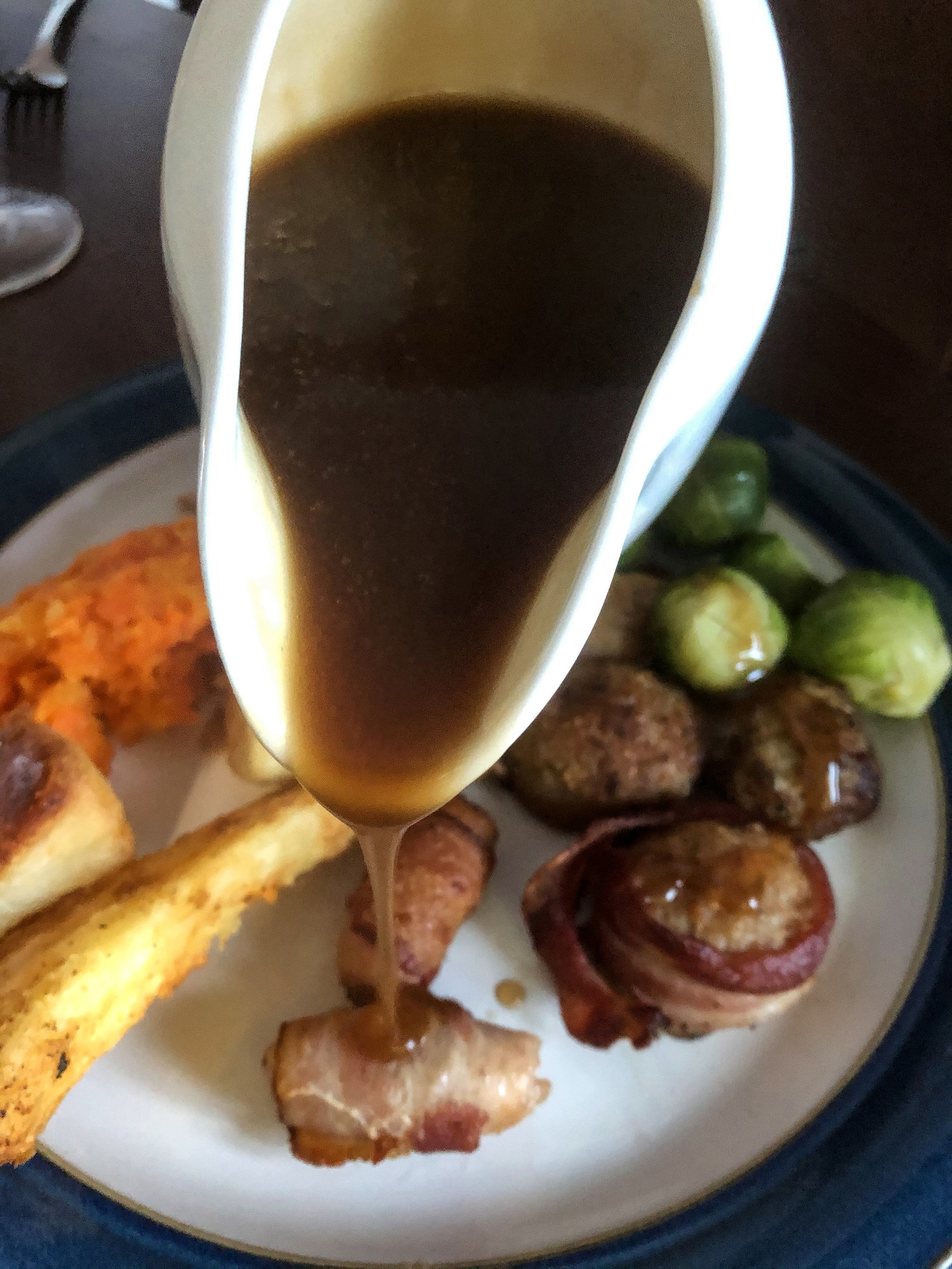 White gravy jug pouring over roast dinner of turkey, carrot and turnip mash, pigs in blankets, pork meatballs, Brussels sprouts, roast potatoes, and parsnips