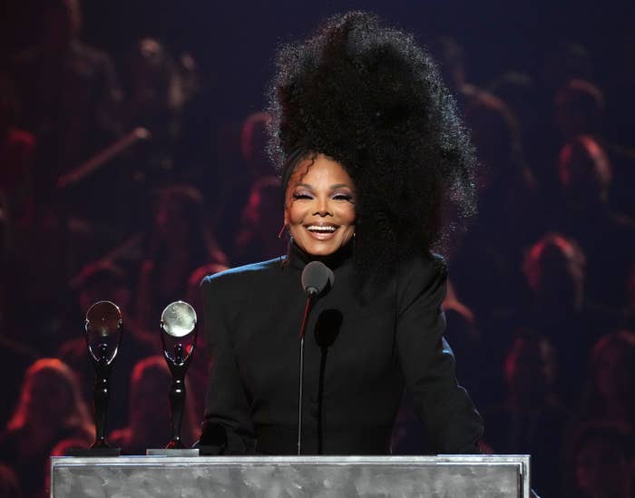 Janet at a podium, smiling widely. She wears all black and her hair is worn on one side