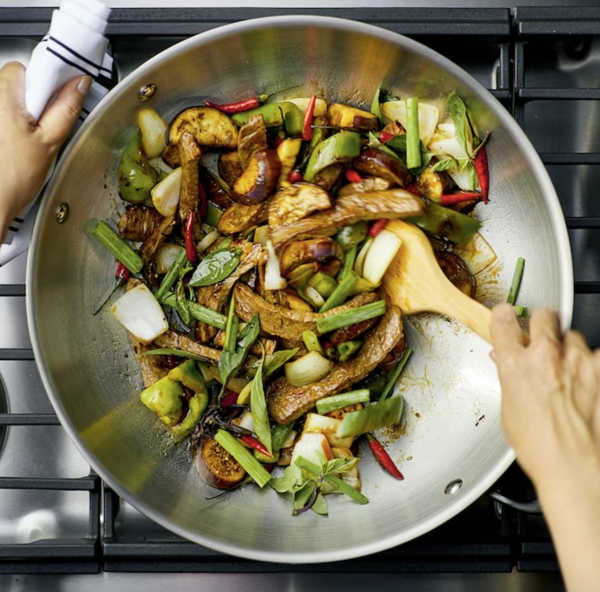 a person cooking vegetables with a wok and wooden spoon over a stove top