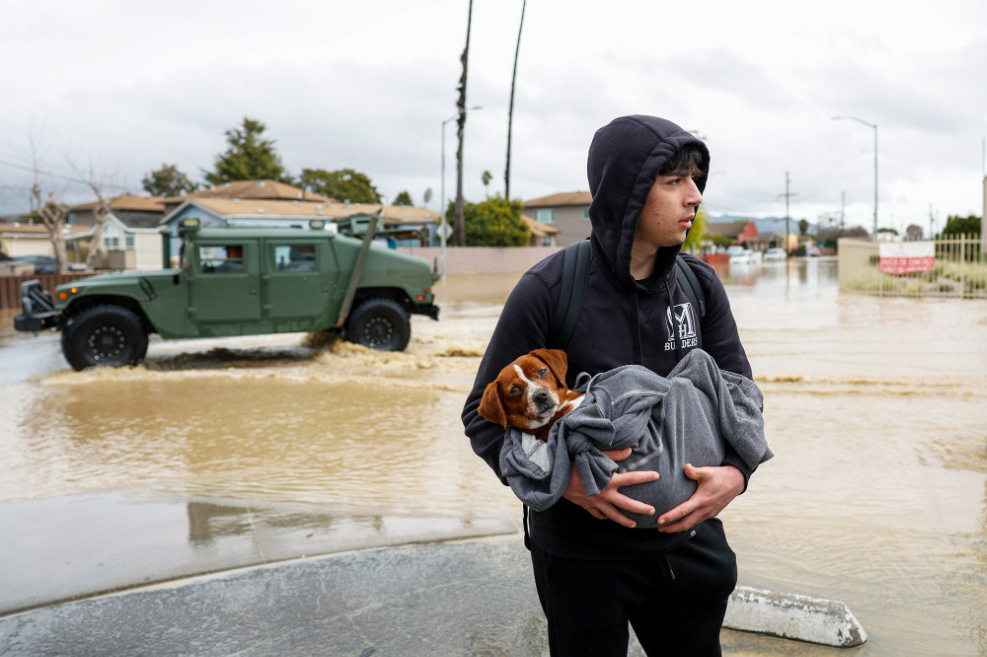 A teen holds a dog wrapped in a blanket as he stands in a flooded neighborhood