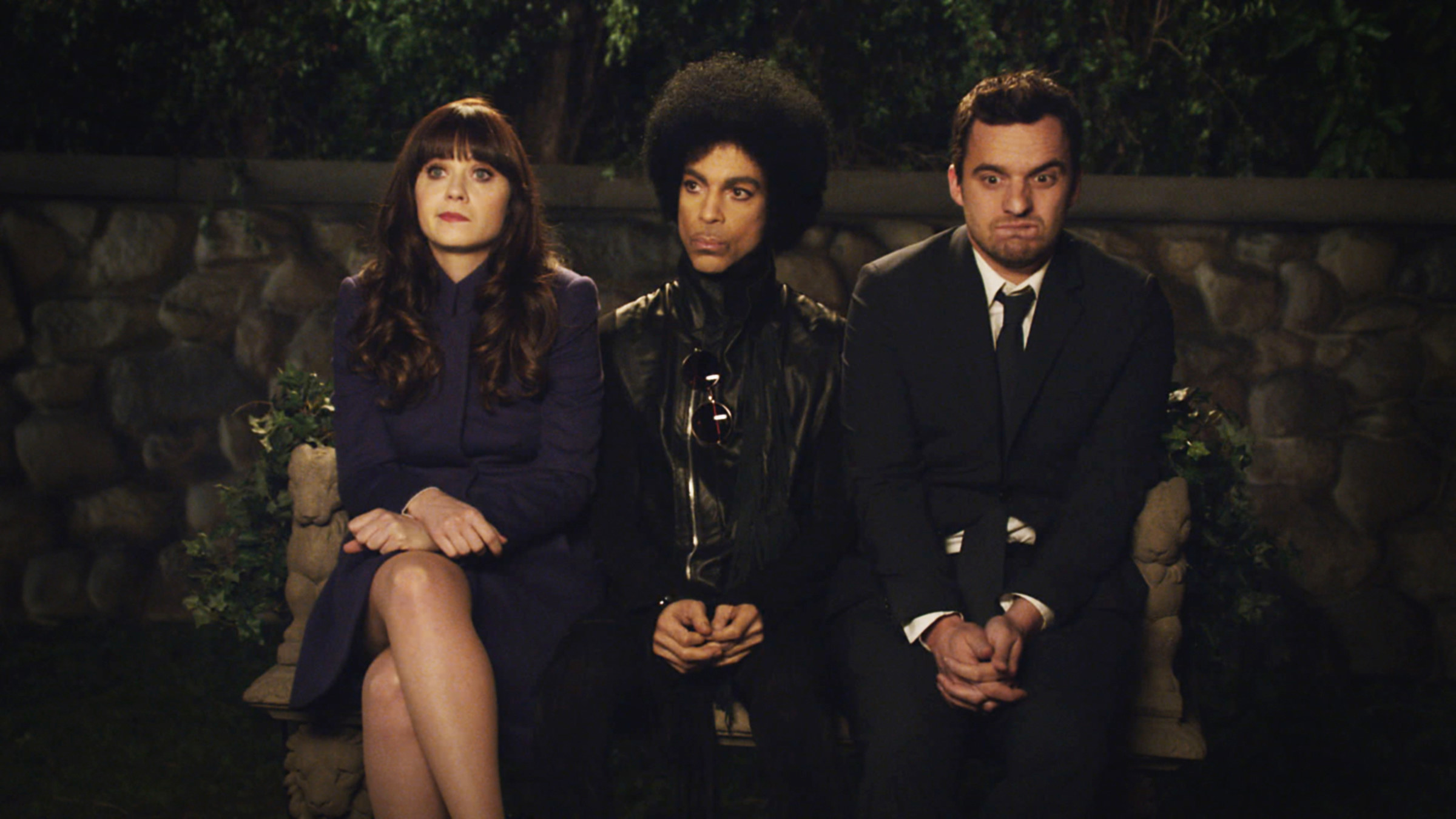 Zooey Deschanel, Prince, and Jake Johnson sitting together on a bench, looking forward with serious expressions. Deschanel in a blue dress, Prince in a black outfit with a shiny blouse, Johnson in a suit