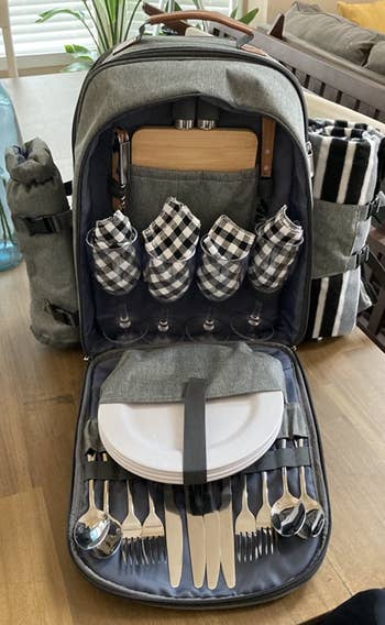 The picnic backpack, opened to show lots of compartments full of plates, silverware, napkins, a cutting board, picnic blanket, and more
