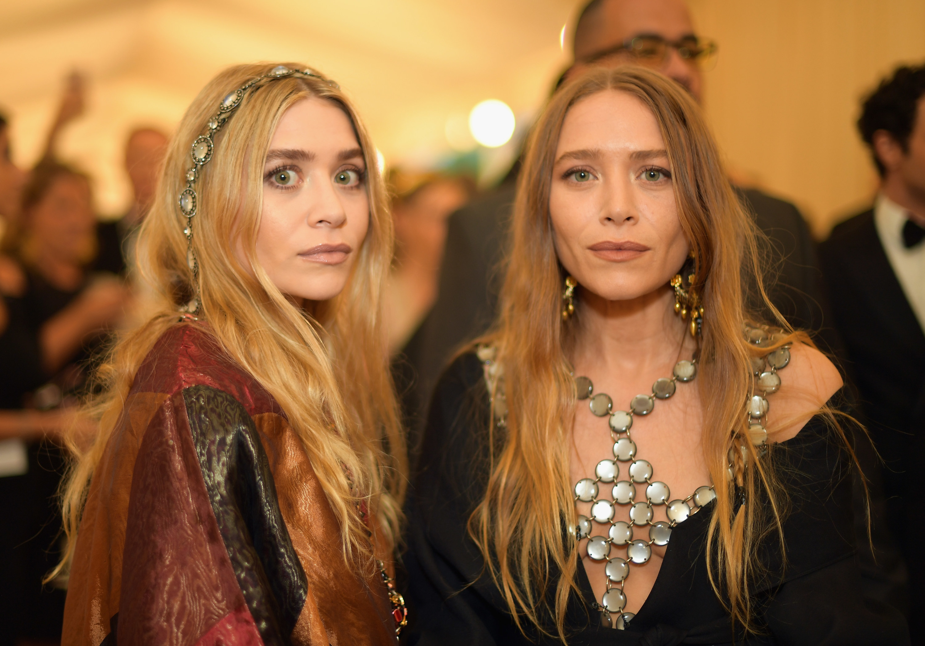 Ashley wearing a medallion style head band with a bohemian style dress, next to Mary Kate with a chest piece made of circular beads with a black dress