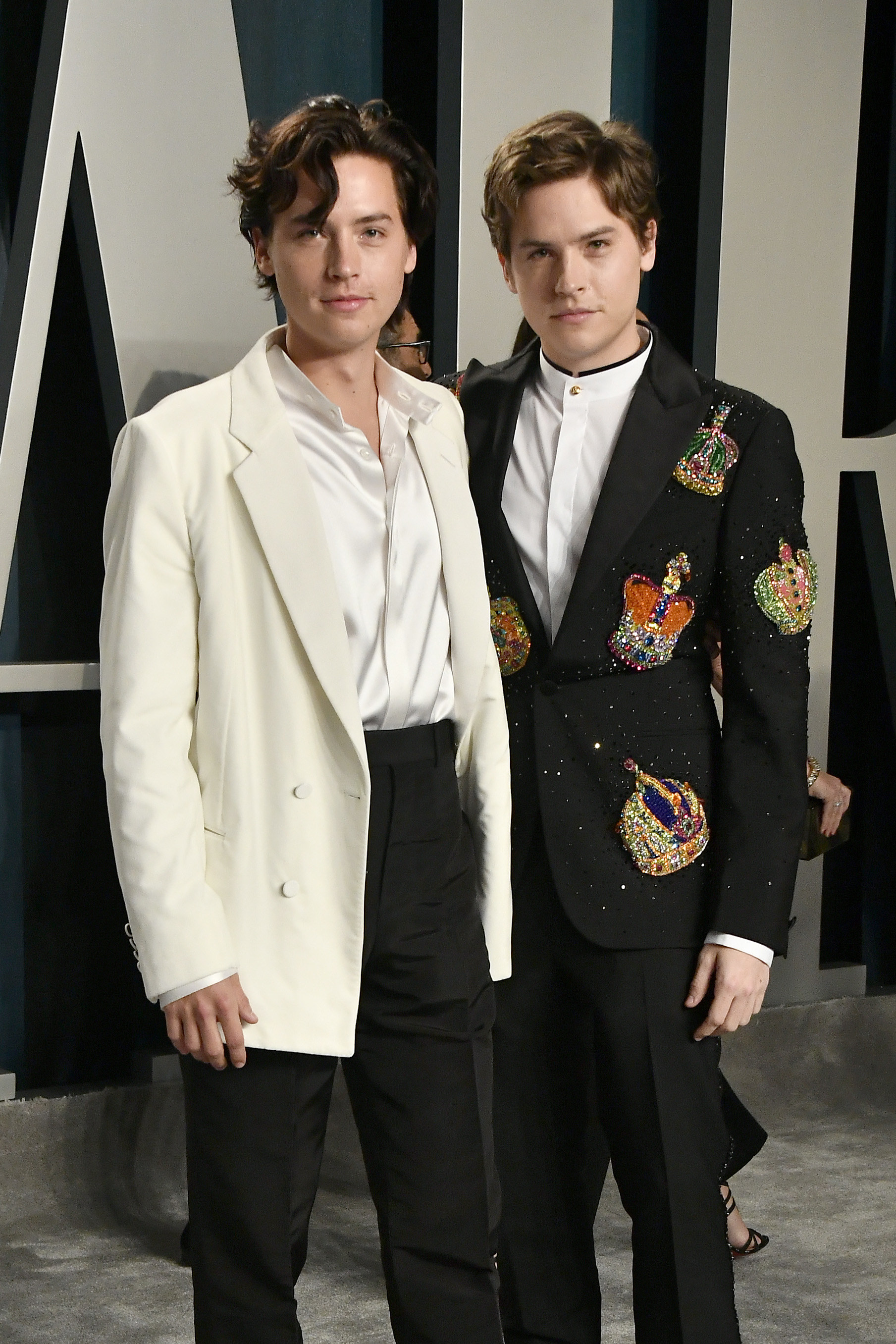 Cole and Dylan at an event. Cole wears an oversized off-white blazer with white shirt and black trousers, while Dylan wears a statement black blazer with embroidered crowns on it