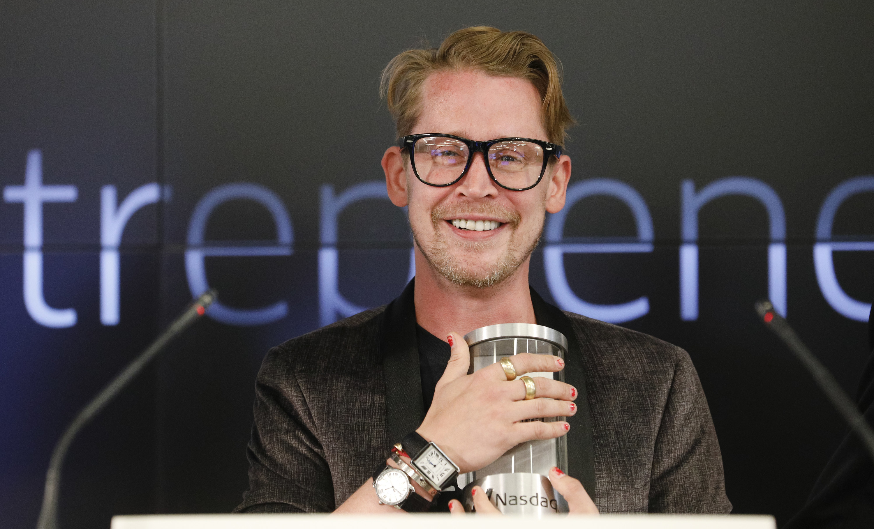 Macauley wearing large black rimmed glasses, and smiling widely while holding a capsule of some sort