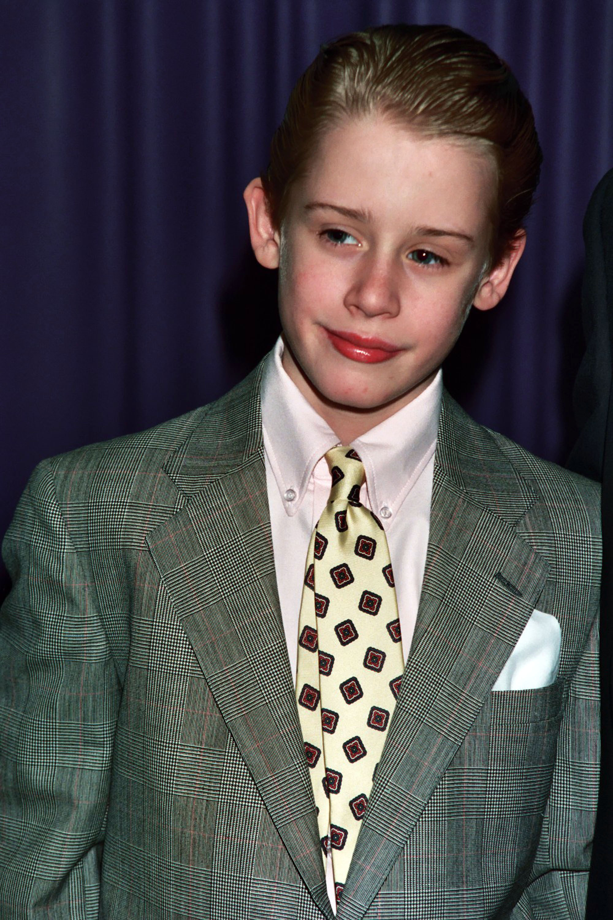 A photo of Macaulay as a kid, wearing a gray tweed suit jacket with a pale yellow tie with red and black squares on it
