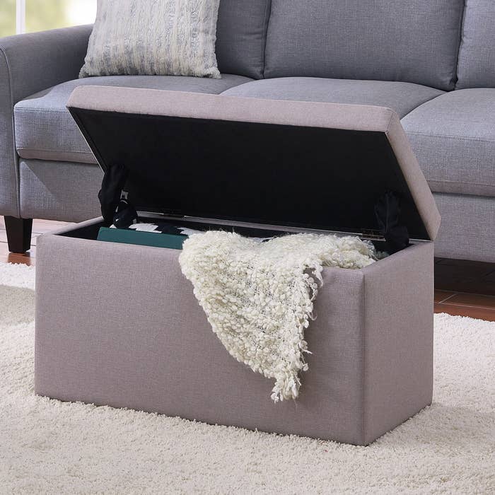 a storage ottoman in sand holding a blanket next to a sofa