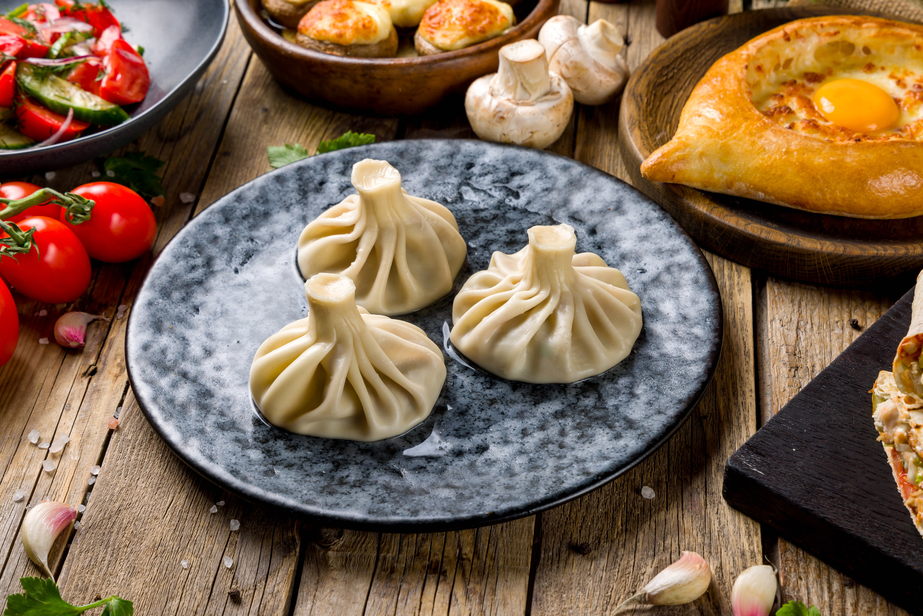 A plate of khinkali on a table along with other Georgian dishes such as khachapuri and tomatoes and cucumbers